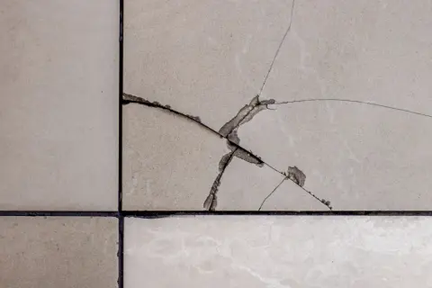 Gros plan sur un carrelage de sol beige avec une fissure importante. Il faut réparer cette fissure pour éviter qu'elle ne s'aggrave.