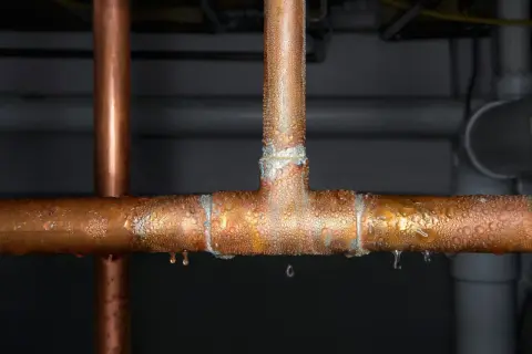 Gouttes d'eau sur un raccord en T de tuyau de cuivre, signe d'une fuite. L'eau perle sur le métal, certaines gouttes tombent.