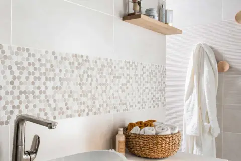 Détail d'une salle de bain moderne avec une bande de pose de faïence en mosaïque hexagonale. Un panier de serviettes et un peignoir blanc complètent la scène.
