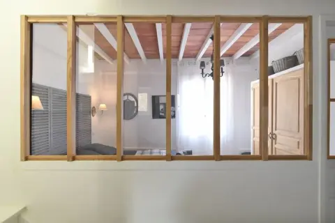 Une verrière en bois sépare deux pièces. On voit des volets, un lit, un lustre et des portes. Idéal pour savoir comment installer une verrière.