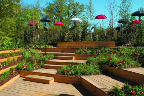 Escalier extérieur en bois avec des parterres de fleurs et des parasols colorés. La norme hauteur marche escalier extérieur est respectée pour un accès sûr.