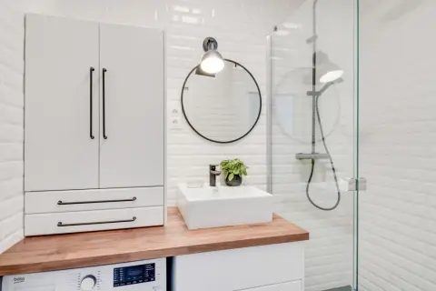 Coin buanderie dans salle de bain moderne avec machine à laver, lavabo, miroir rond et douche vitrée.