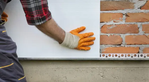 Pose d'isolant blanc sur un mur de briques. La main gantée ajuste la plaque, une étape clé dans la définition de l'isolation.