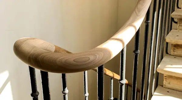 Courbe élégante d'une main courante en bois sur une volée d'escalier aux barreaux de fer forgé, baignée de lumière.