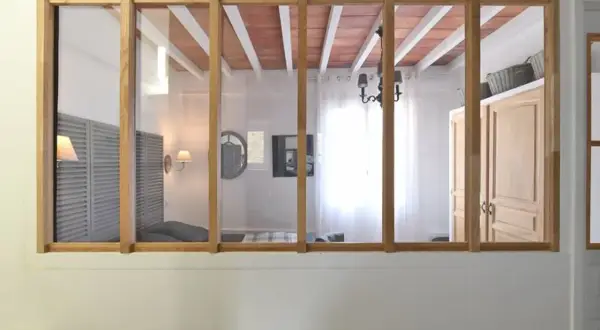 Une verrière en bois sépare deux pièces. On voit des volets, un lit, un lustre et des portes. Idéal pour savoir comment installer une verrière.