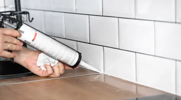 Application d'un joint d'étanchéité blanc sur un plan de travail en bois, près de carrelages muraux blancs. Le temps de séchage de la colle carrelage flex est une préoccupation pour une finition parfaite.