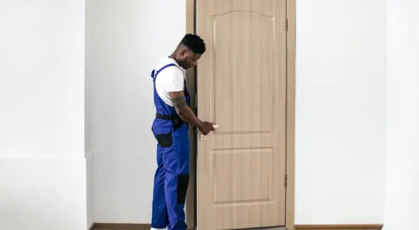 Un ouvrier en salopette bleue manipule la poignée d'une porte intérieure en bois clair, prêt à la changer.