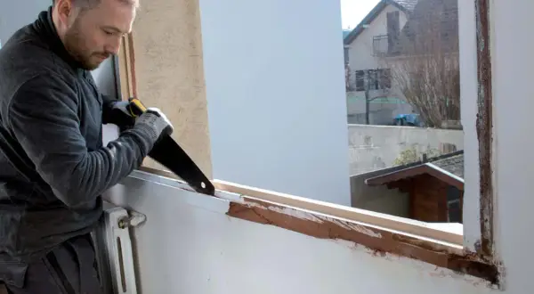 Un bricoleur prépare le cadre pour comment poser une fenêtre PVC en rénovation, sciant le bois existant.