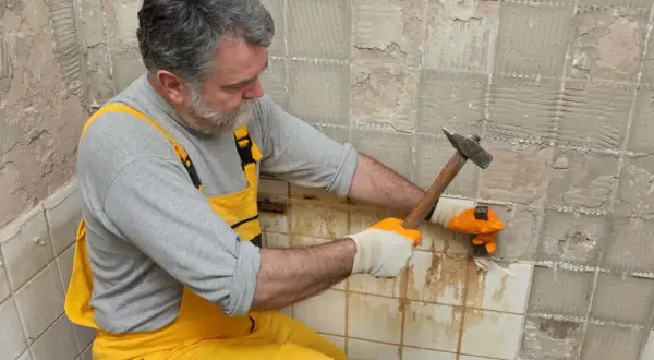 Homme en salopette jaune en train d'enlever du carrelage mural avec un marteau et un burin.