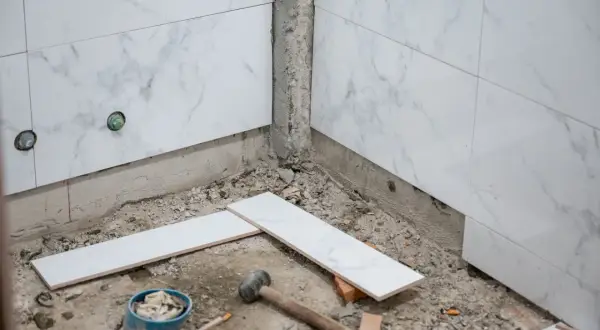 Début des travaux pour carreler une salle de bain : des carreaux de marbre blanc sont posés sur le sol, près d'un marteau et d'un seau.